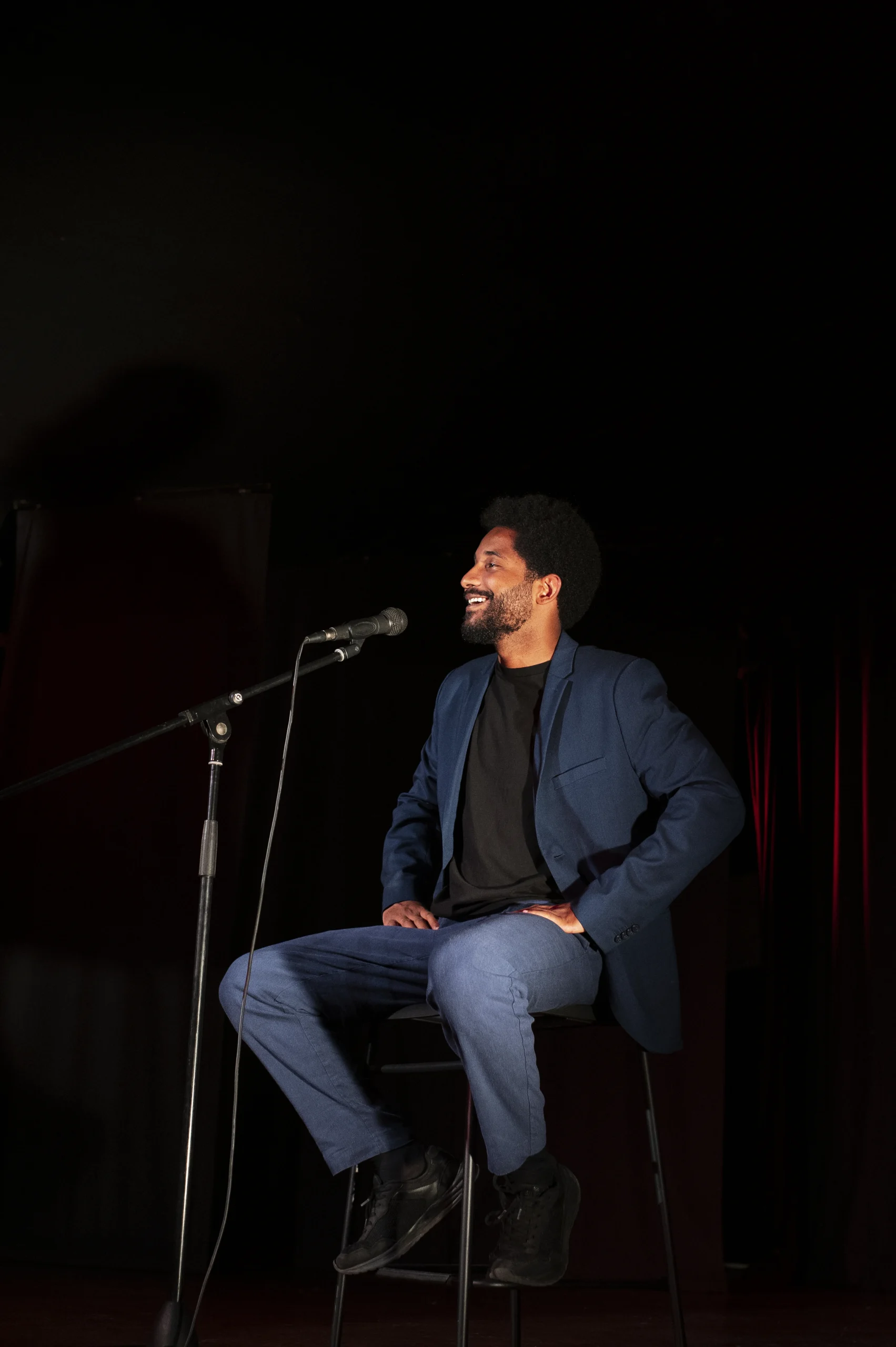 Man in blue blazer seated on stool on stage, speaking into microphone during a stand-up comedy performance
